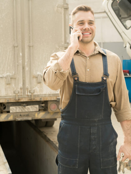 Auto technician standing in service garage with broken trucks, talking on smartphone and smiling cheerfully