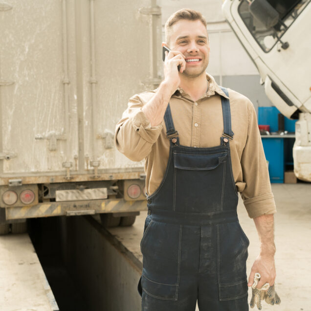 Auto technician standing in service garage with broken trucks, talking on smartphone and smiling cheerfully