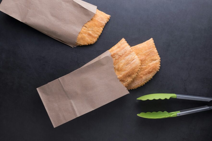 Aerial view of three Jamaican beef patties in brown paper bags with a tong on a black background